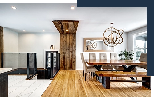 photo of a bright, modern loft space: rectangular marble kitchen tiles, sleek wrought iron railing, wooden flooring with skinny boards, a large wooden trestle table with wrought iron cross-bars in the kitchen space plus a matching bench and five high-backed upholstered chairs; a custom-designed light fixture with concentric circles like electron paths hangs above the table