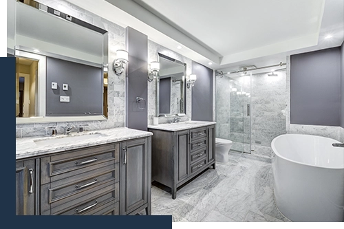 photo of a high-end bathroom renovation: dark grey brown wood vanities with marble counters, grey marbled tiles on the floor and in the glass-doored shower, and a large, sleek soaking tub