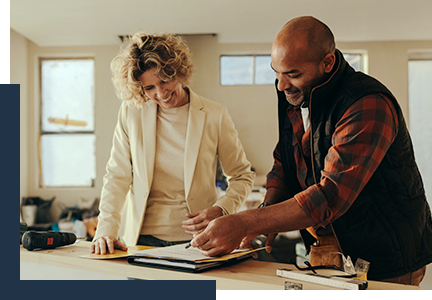 photo of a male contractor in a red flannel shirt and a vest walking through project information with a female homeowner in a cream sweater with a cream blazer