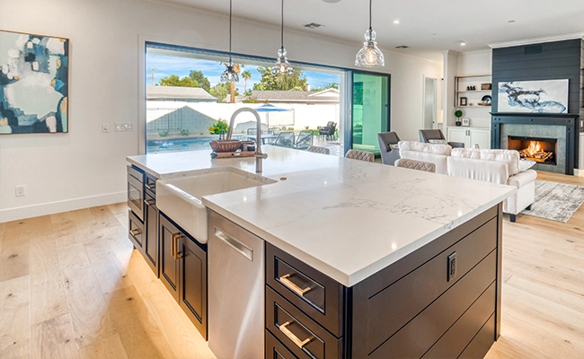 photo of a high-end kitchen featuring a dark wood island with brass drawer fixtures, a farmhouse sink, and a marble countertop; the rest of the space is open concept with blond wooden floors, a huge picture window with sliding glass door, a fireplace, and built-in shelving